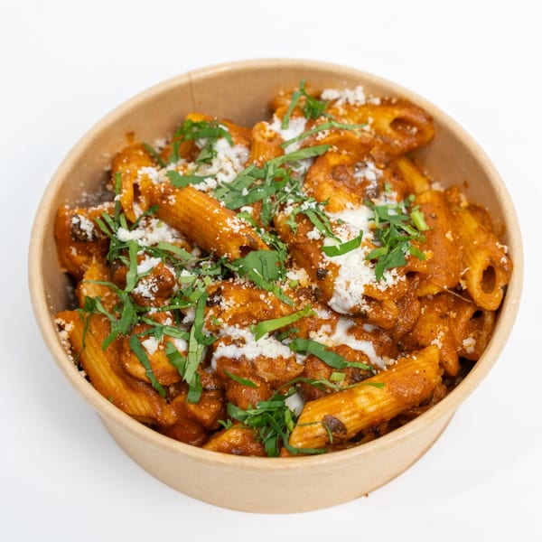 Penne pasta in a rich red sauce, topped with white cheese and fresh green herbs in a brown bowl.