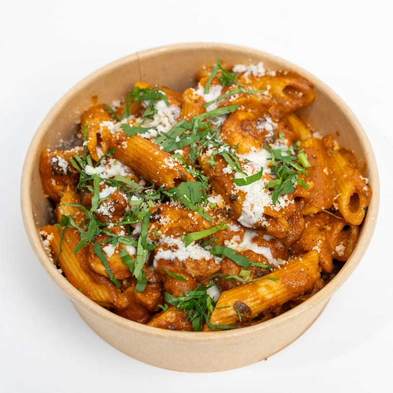 Penne pasta in a rich red sauce, topped with white cheese and fresh green herbs in a brown bowl.