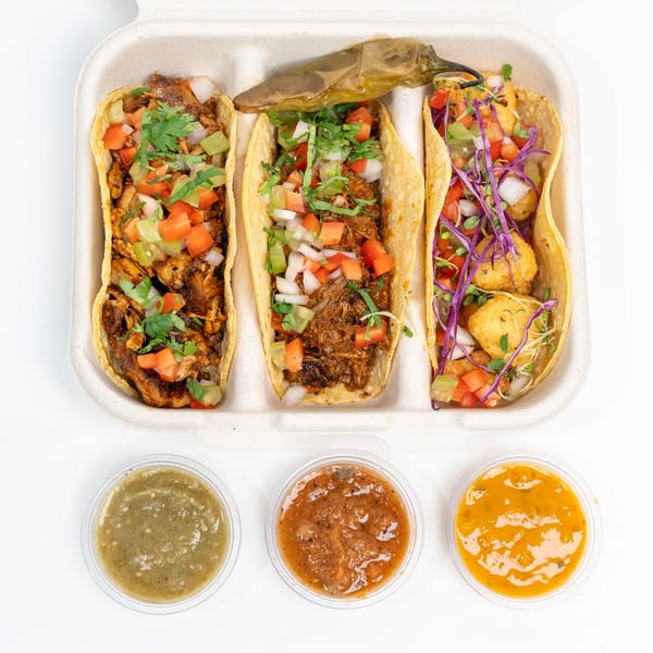 Three Indian-style tacos with various fillings, toppings, and three sauces in a takeout container.